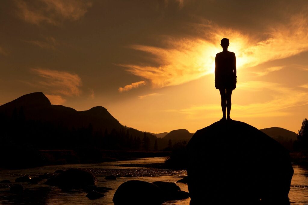 A person standing on top of a rock in front of the sun.